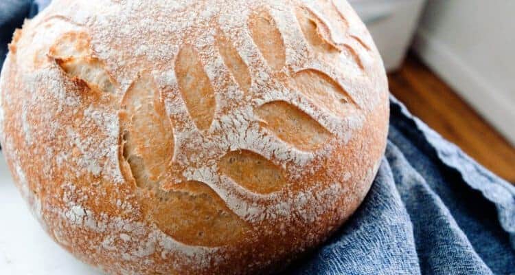 no knead sourdough bread loaf with leaf design on a white countertop with a dark blue towel