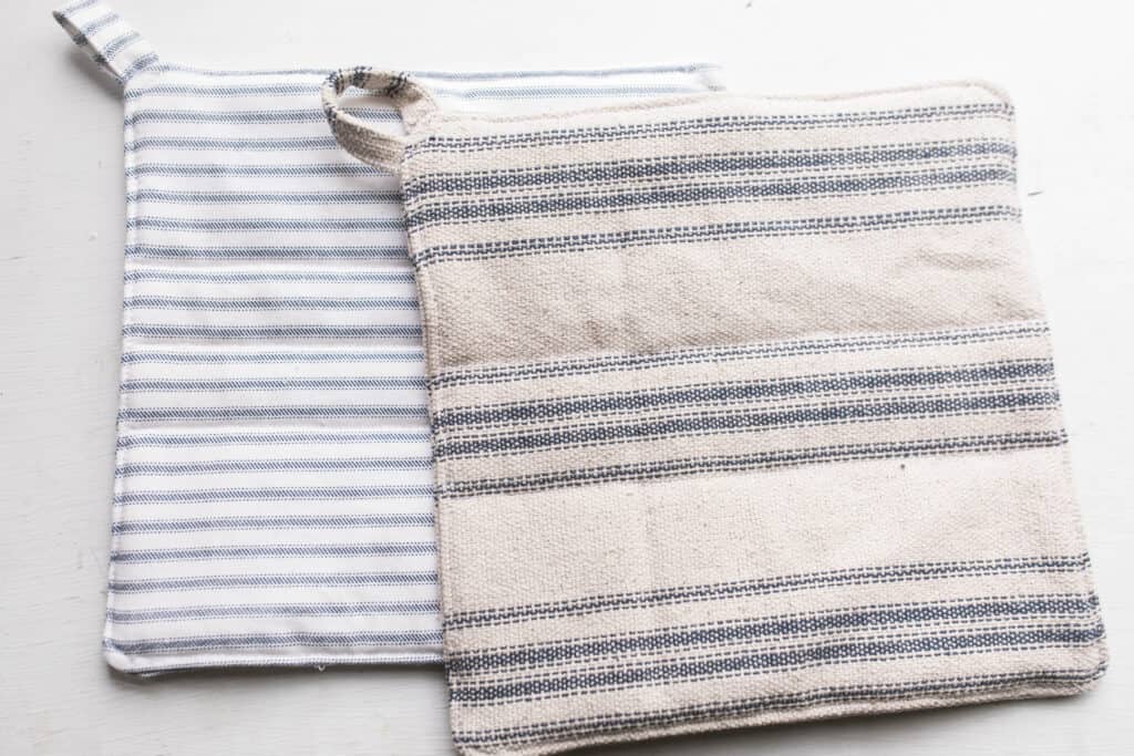 Two homemade pot holders. One blue and white stripped lays on a white table with a blue and cream ticking stripe pot holder on top