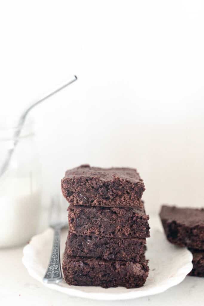 four einkorn brownies stacked on top of each other on a white plate with a mason jar of milk with a stainless straw and a pan of brownies in the background