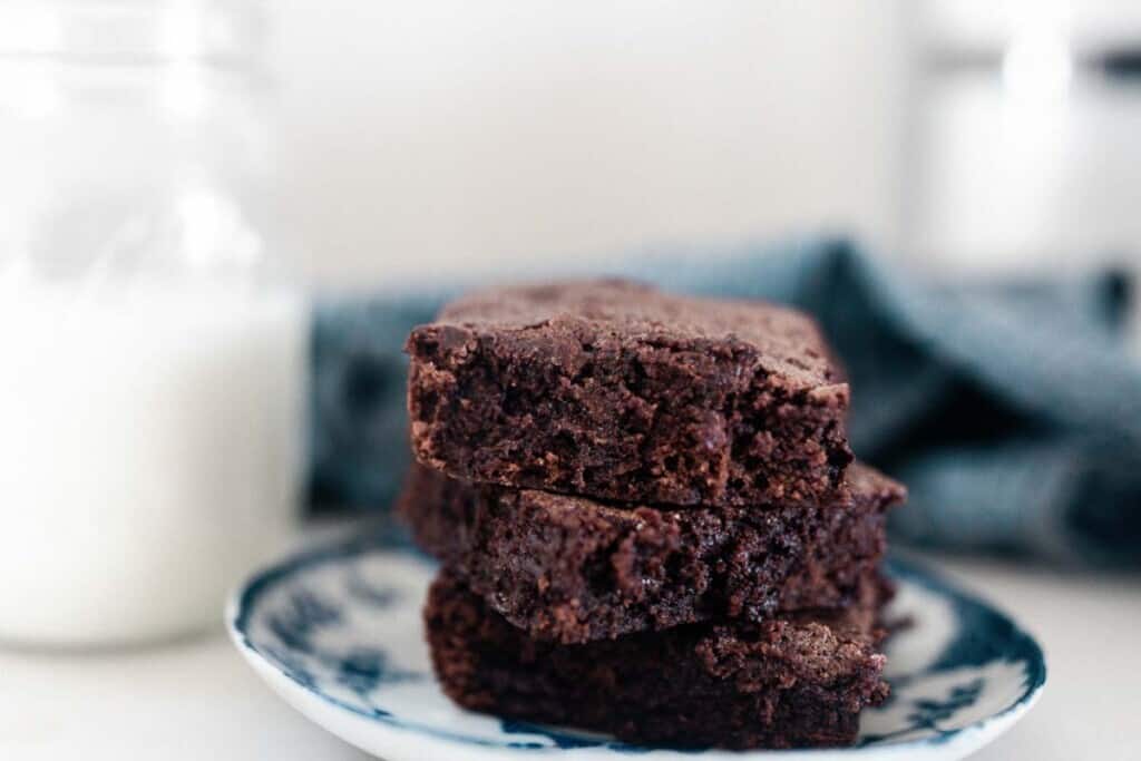 three einkorn brownies stacked on a white and blue antique plate with a mason jar of milk and a blue towel in the background