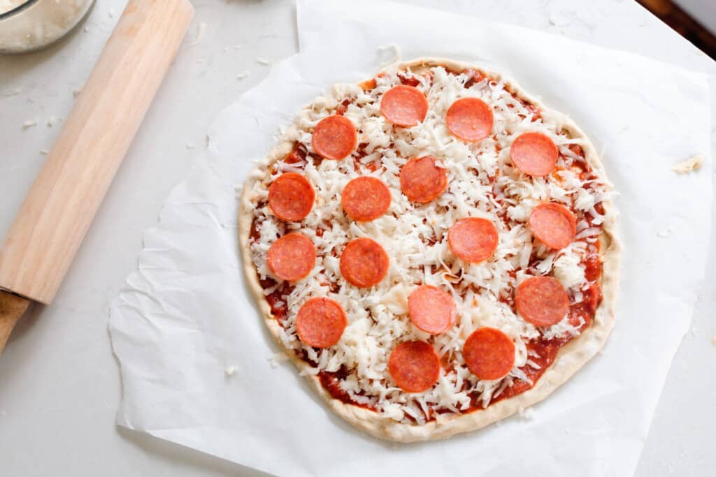 einkorn pizza crust with cheese and pepperoni on parchment paper with a rolling pin to the left