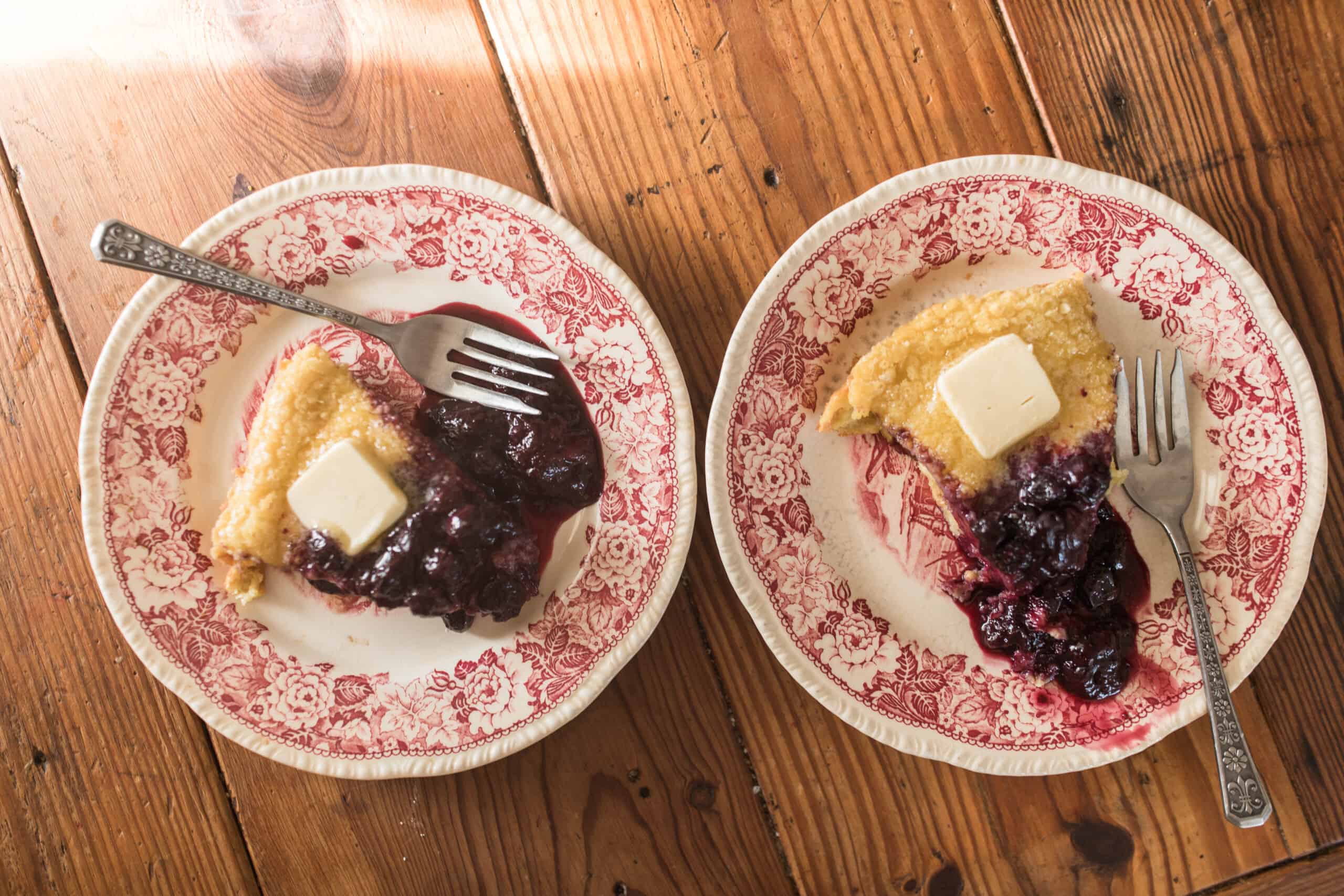 Overhead photo of two vintage plates with einkorn puff pancakes topped with berry syrup with forks on a wooden table.