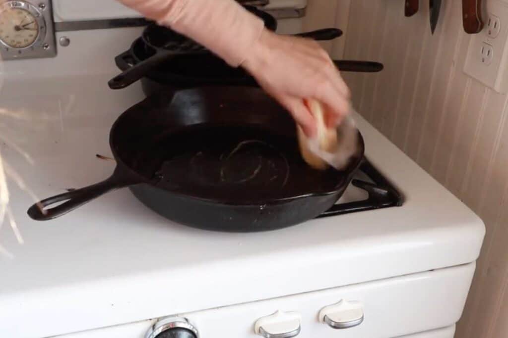 A hand buttering a large cast iron skillet on a stovetop.
