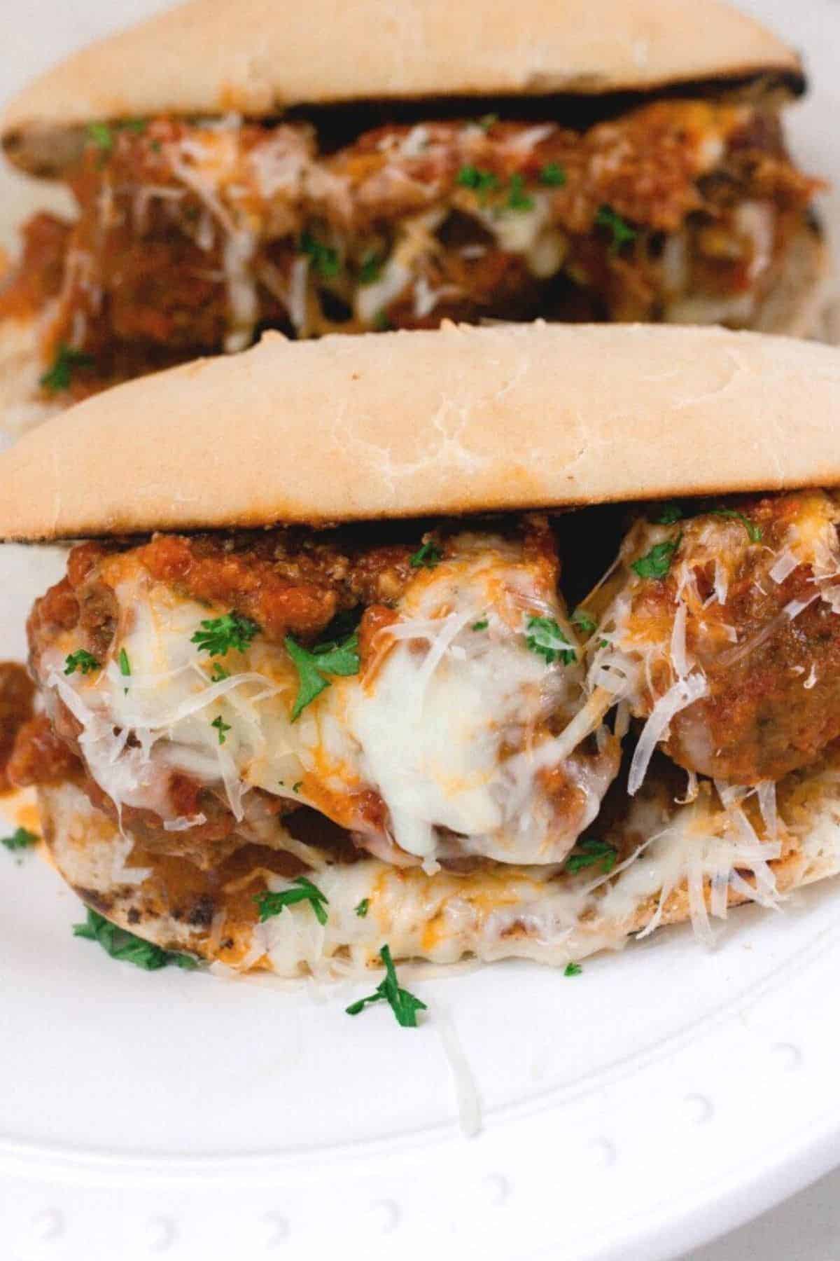 homemade meatball sub sandwich with cheese, marinara sauce, and parsley on a white plate. Another sandwich is in the background