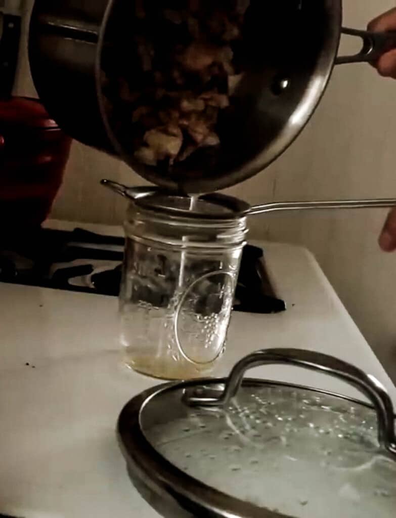 a pot of rendered beef fat being strained through a fine mesh strainer into a mason jar. The pot lid sits right in front of the jar