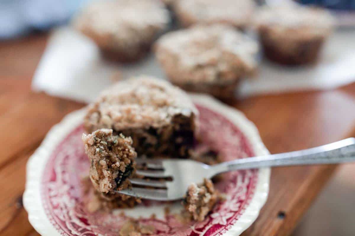 five sourdough discard blueberry muffins on a wire cooling rack on a white countertop with fresh blueberries sprinkled around. The muffin at the corner has the cupcake liner peeled down
