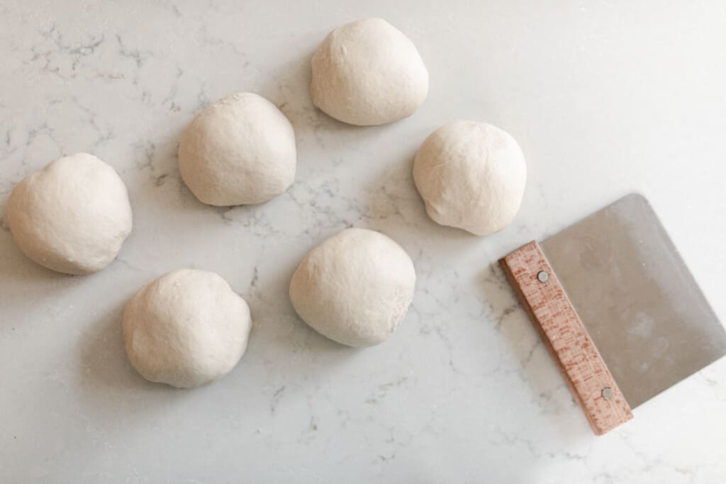 sourdough sandwich roll dough in 6 dough balls on a white countertop with a bench scraper to the left