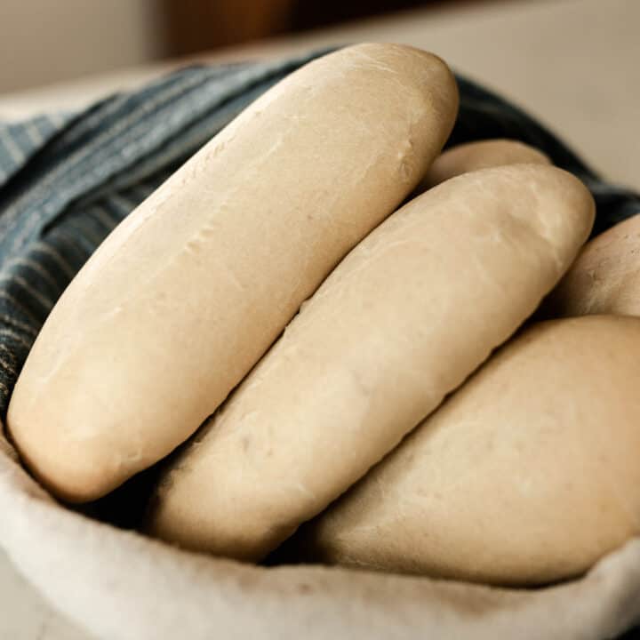 sourdough sandwich rolls in a basket with a blue and white stripped towel on the side
