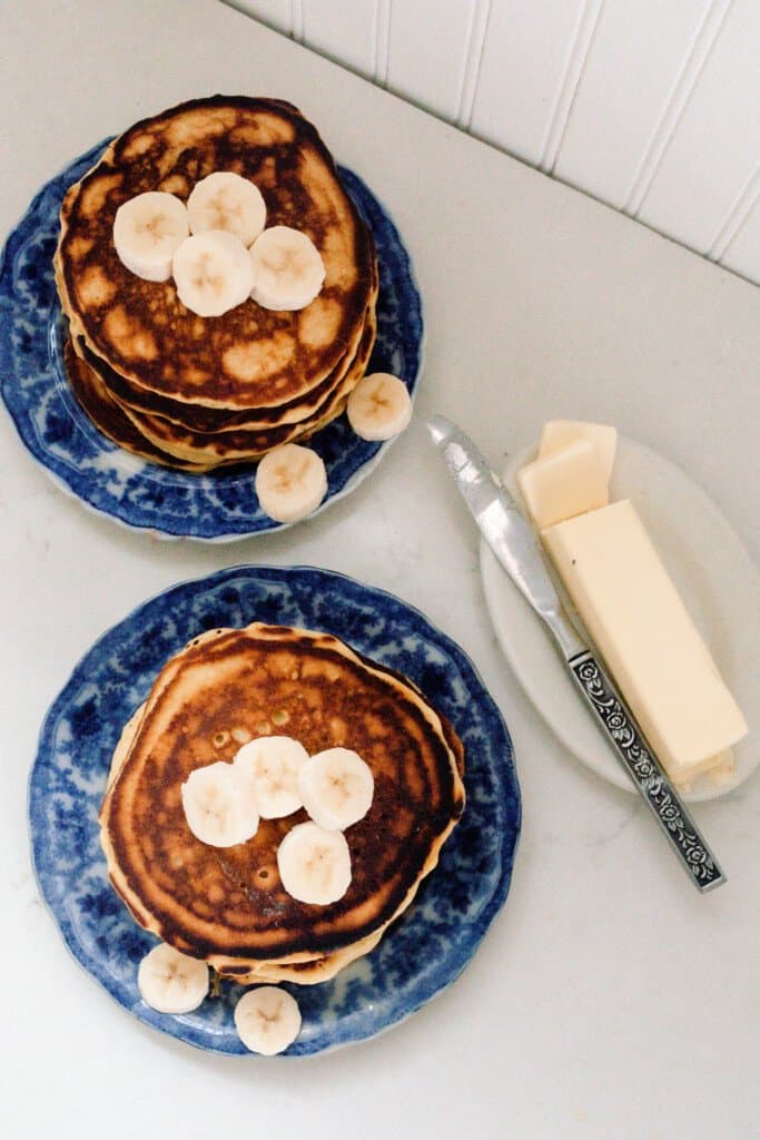 overhead photo of einkorn pancakes topped with bananas on a blue and white antique plates. a dish with butter and a knife sit to the right of the pancakes