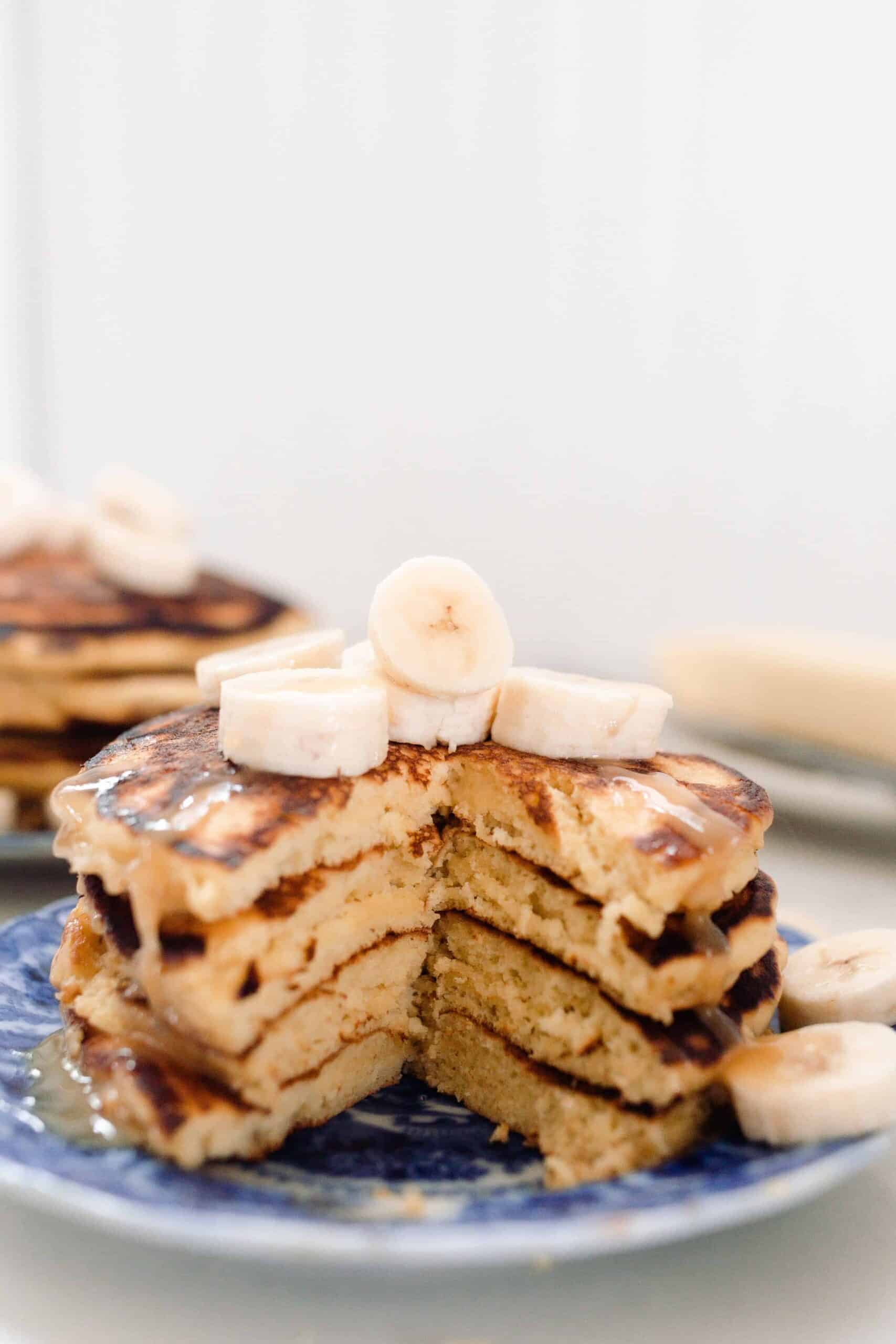 stack of einkorn pancakes topped with bananas on a blue and white antique plate. Butter on a dish is in the background