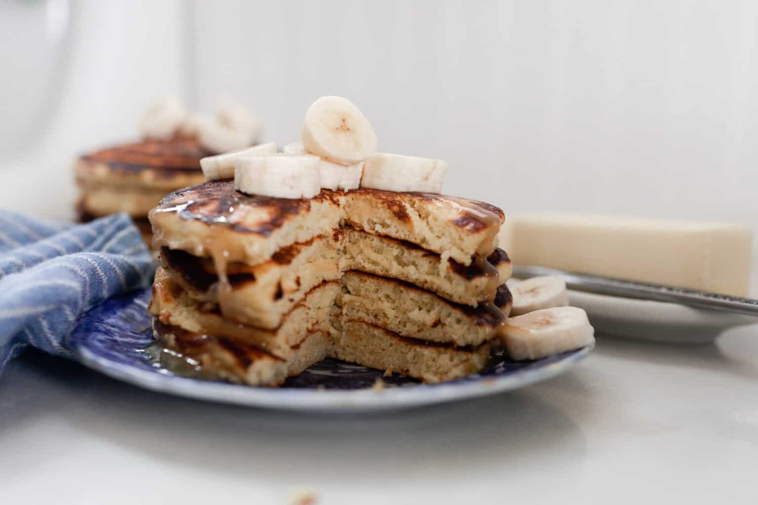 two stacks of einkorn pancakes with bananas on a white and blue antique plate on a white countertop