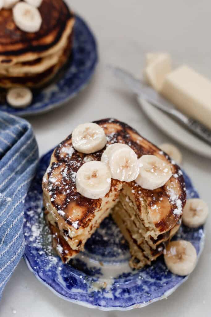 stack of einkorn pancakes topped with bananas with slice taken out of the stack. Butter on a dish with a knife to the right