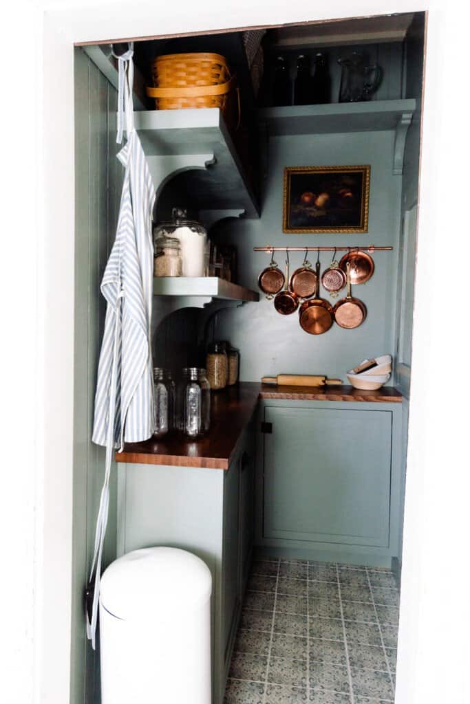 farmhouse pantry reveal with custom blue/green cabinetry, shelving, peach art in a vintage frame, and copper pots hanging on the wall.