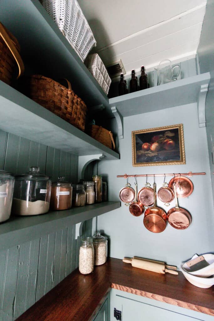 blue green painted shelves with corbels with jars and baskets lining the shelves. copper pots hand on a copper pot hanger with a peach painting in a vintage frame hangs above it.