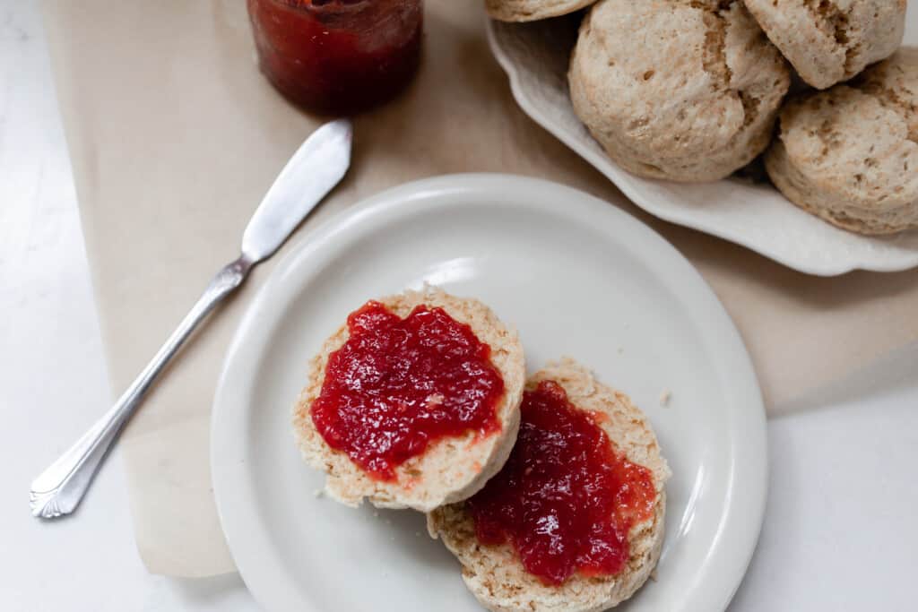 sourdough biscuits sliced in half and covered with strawberry jam on a white plate. The white plate is on a piece of parchment paper with a knife to the right. More biscuits on a white plate sit in the back right corner