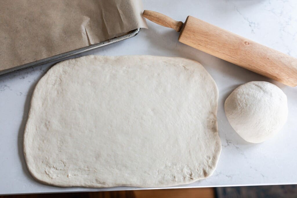sourdough French bread dough rolled out into a square on a white quartz countertop with a rolling pin and more dough to the back right