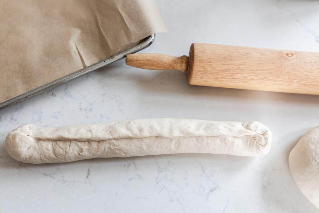 sourdough French bread sough rolled up on a quarts countertop with a baking sheet covered with parchment paper to the left and a rolling pin in the back right
