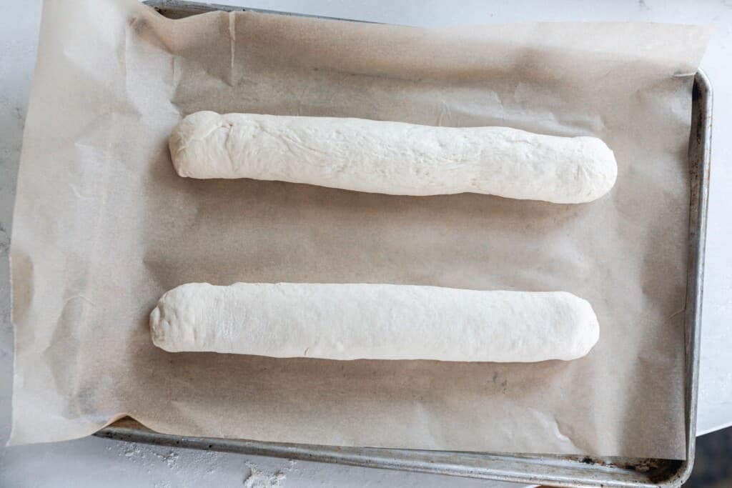 two sourdough baguette dough rolls on a parchment lined baking sheet ready to go into the oven