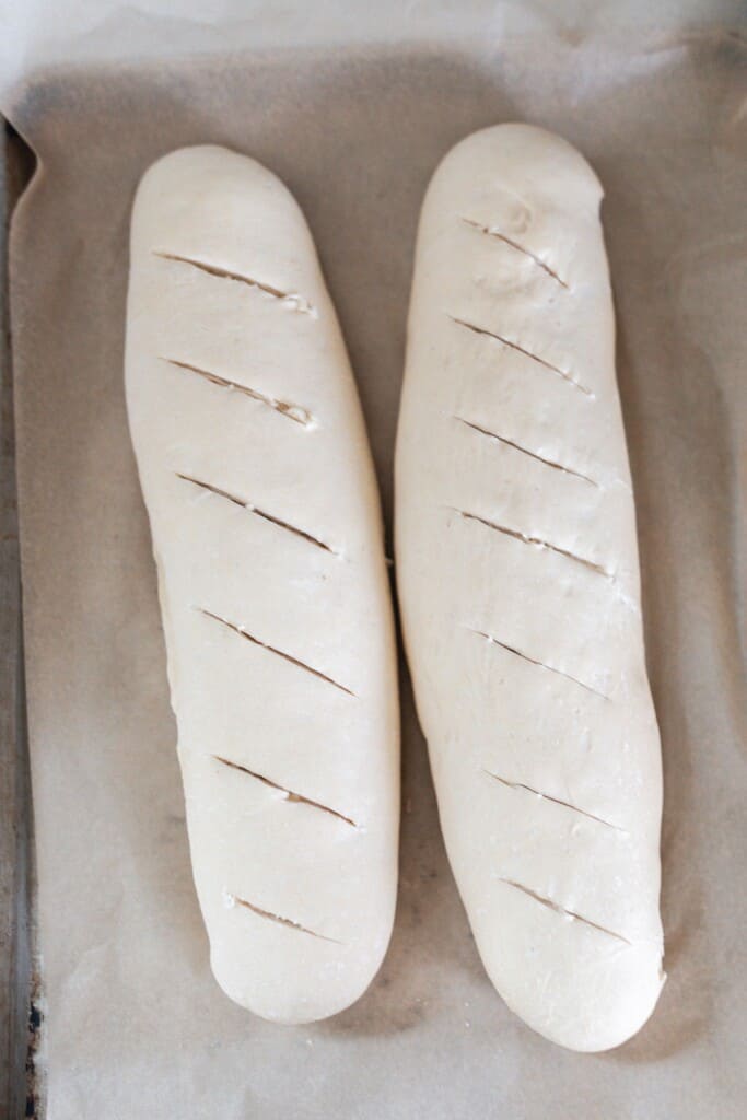 two raw sourdough baguettes slashed with a lame on top of a parchment lined baking sheet ready for the oven