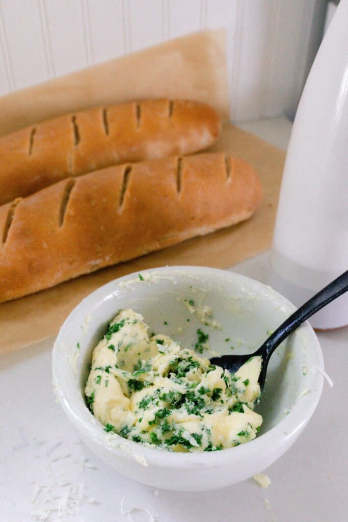 garlic butter in a white bowl with a spoon with two loaves of sourdough French bread straight from the oven on parchment paper