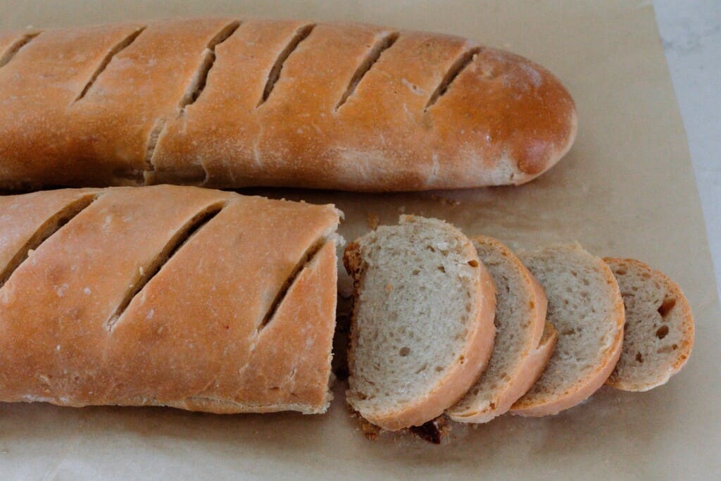 two loaves of sourdough French bread on parchment paper, on is sliced while the other is whole