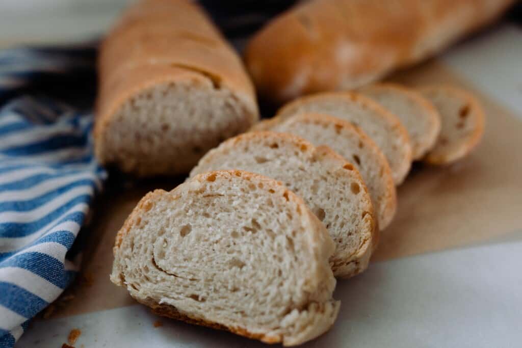 slices of French bread on a wood cutting board with the other baguettes and a blue and white stripped towel in the background