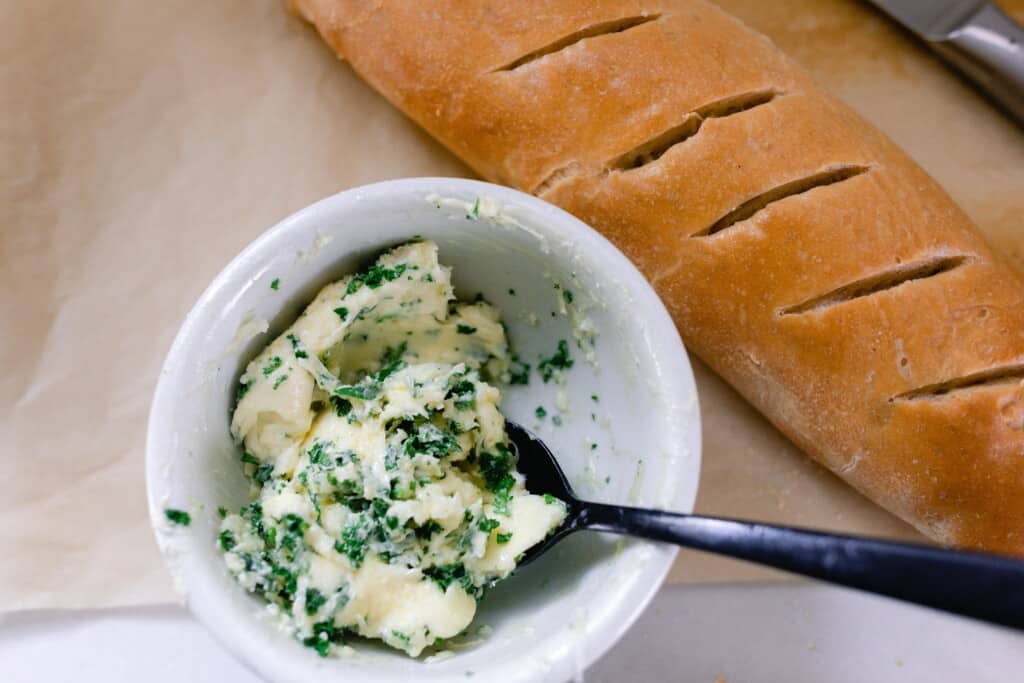 garlic butter in a bowl with herbs next to a loaf of sourdough French bread