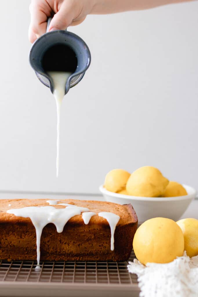 pitcher of lemon glaze being poured over a sourdough lemon pound cake on a wire rack with lemons to the right.