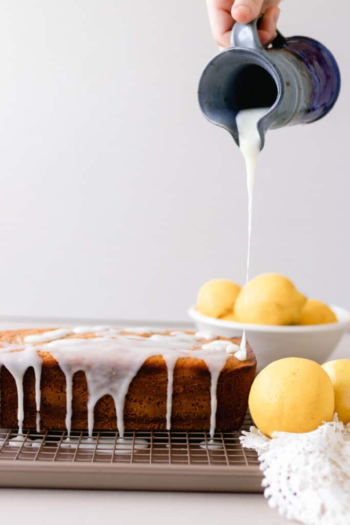 white glaze being poured over a sourdough pound cake on a white rack with lemons to the right in white bowls