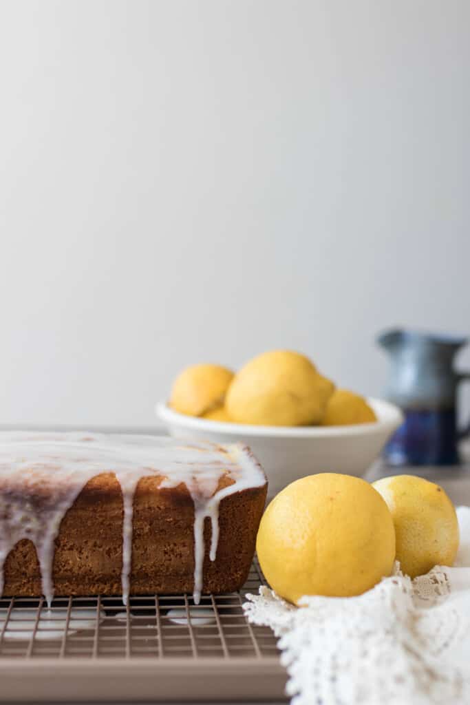 sourdough lemon pound cake drizzled with lemon icing on a wire rack. A bowl of lemons sit to the right