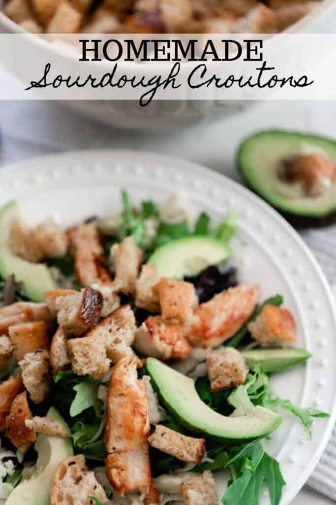 a white plate with a greens, avocados, and sourdough croutons with a bowl of croutons and a half of avocado in the background