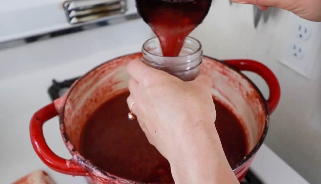 women pouring strawberry jam into a mason jar over a pot of strawberry jam
