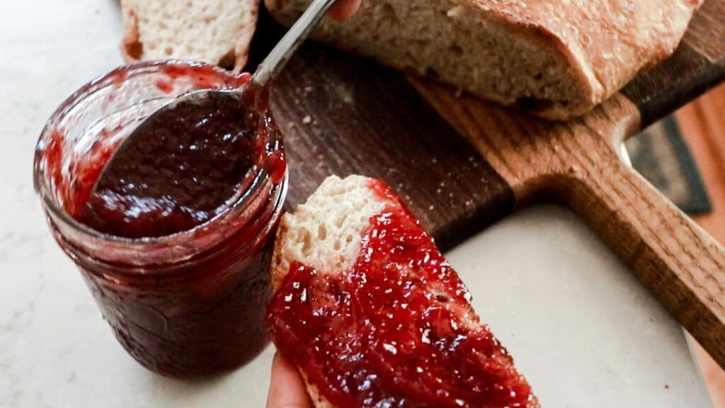 slice of homemade bread covered in jam with jam in a jar behind the bread.