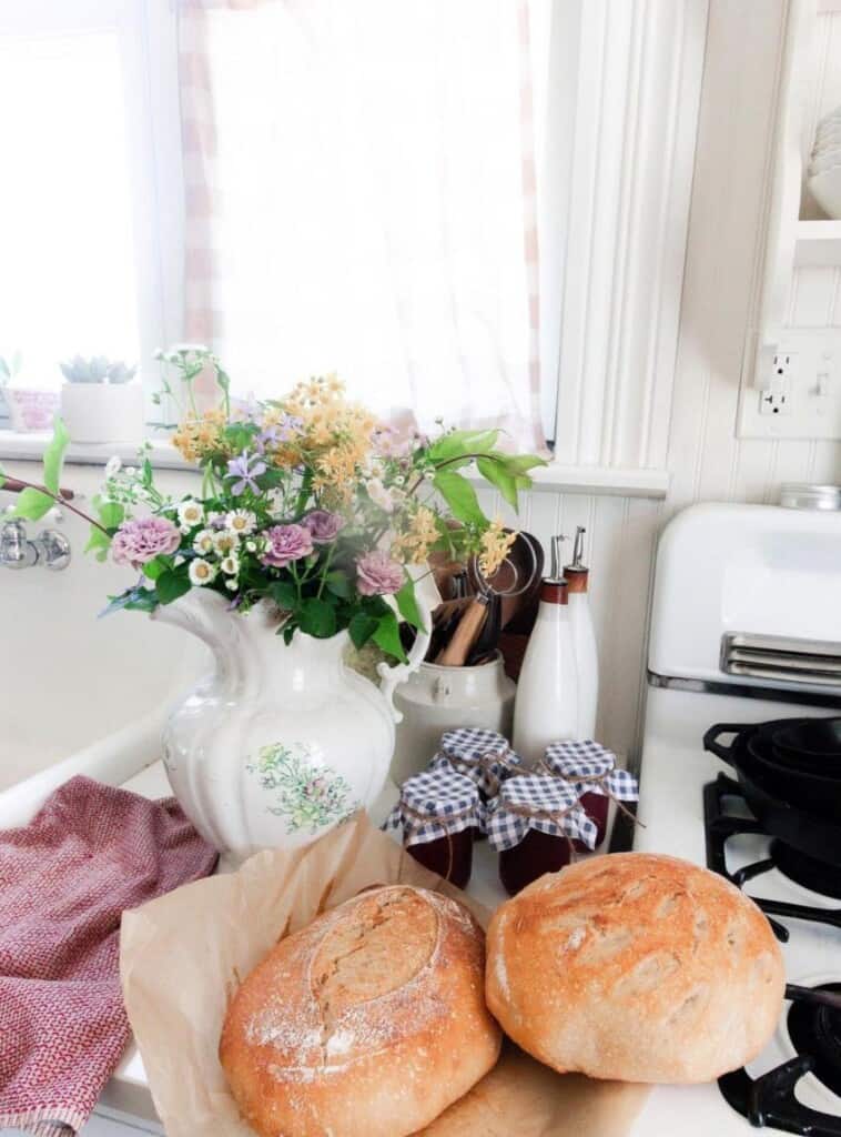 two loaves of homemade bread on parchment paper with a bouquet of flowers in an antique vase with two jars of jam to the right