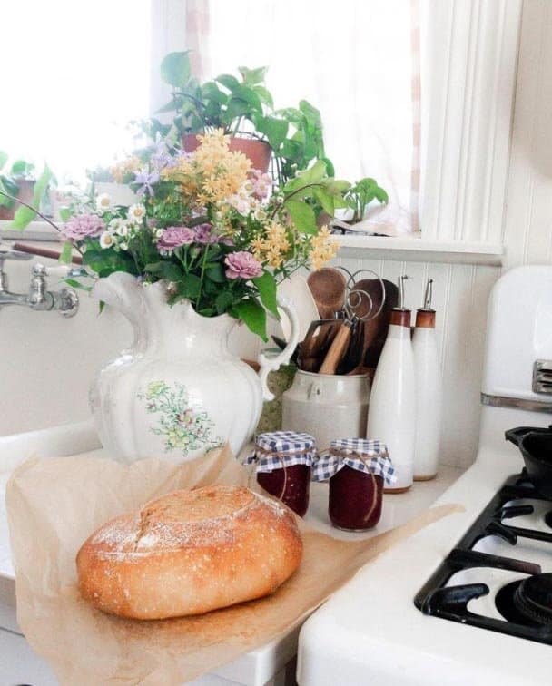 a loaf of homemade sourdough bread on parchment paper on a white countertop with two jars of jam and a bouquet of flowers in a vase in the background