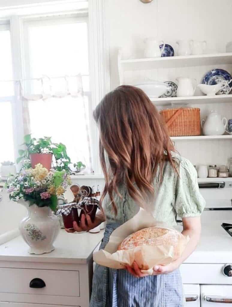 women holding a loaf of homemade bread in one hand and a couple jars of strawberry jam in the other hand in a white kitchen