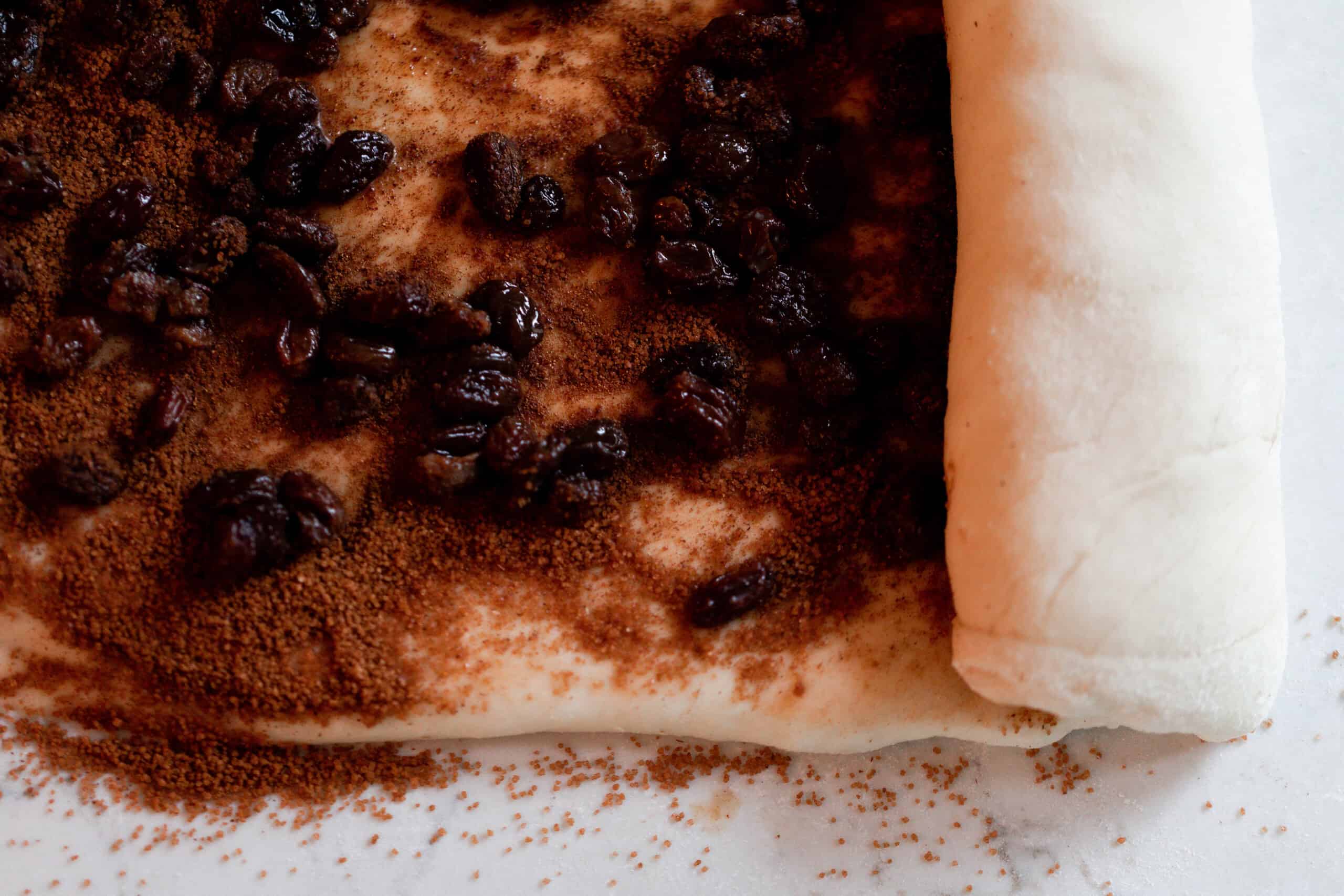 sourdough bread dough with cinnamon, sugar, and raisin sprinkled on the dough being rolled up