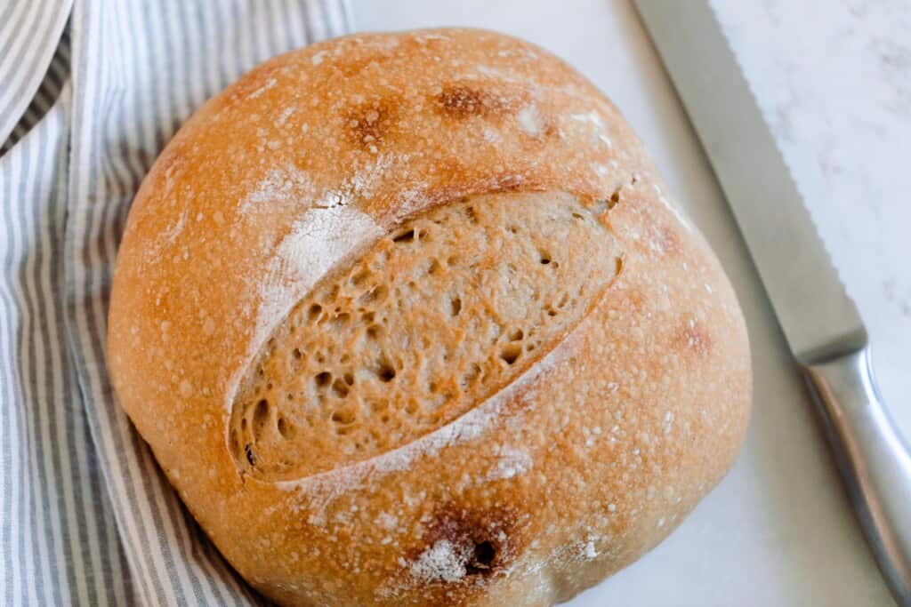 loaf of homemade sourdough bread on a white and light blue stripped towel with a knife to the right