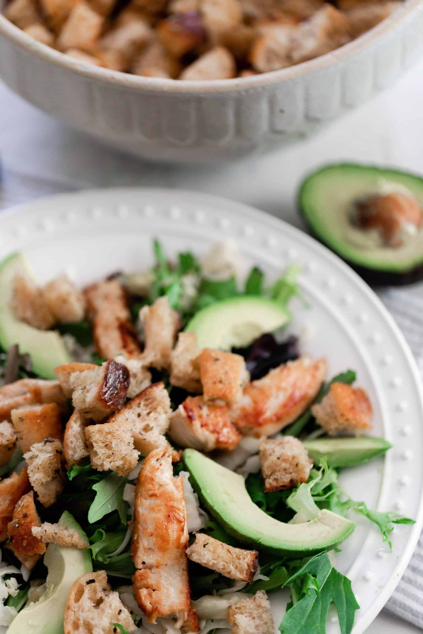 a white plate with a greens, avocados, and sourdough croutons with a bowl of croutons and a half of avocado in the background