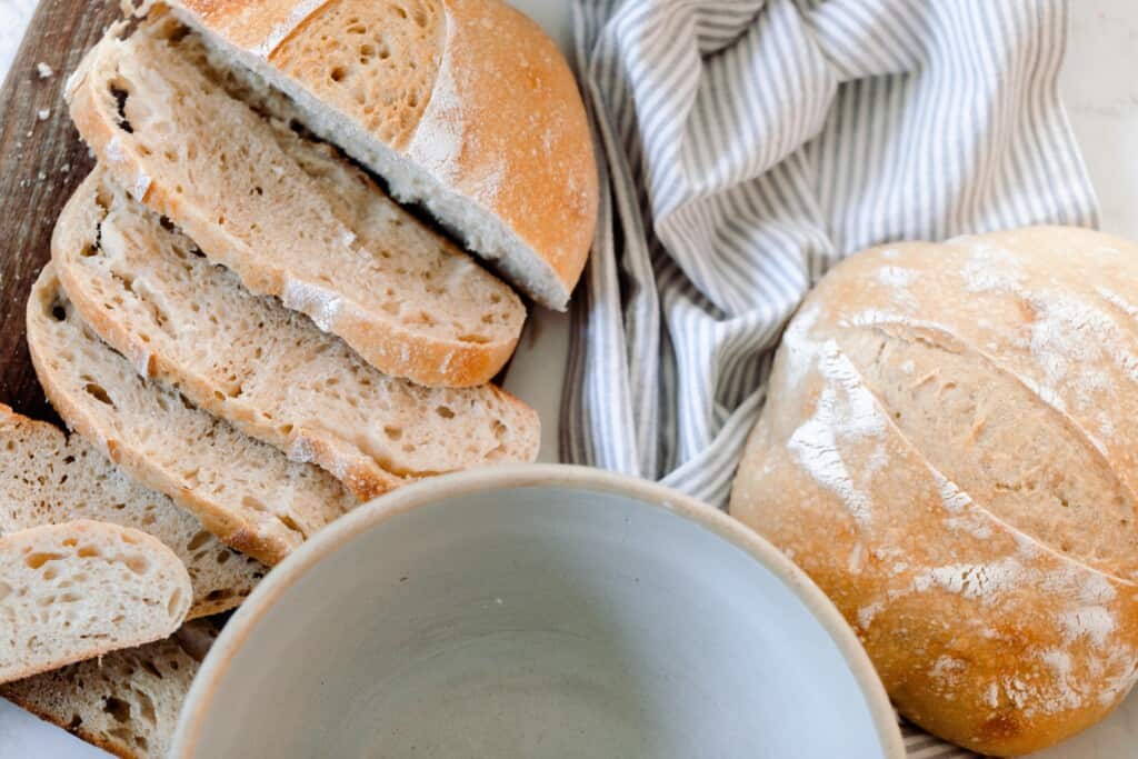 homemade sourdough bread sliced on a cutting board with a large bowl and another loaf of bread to the right