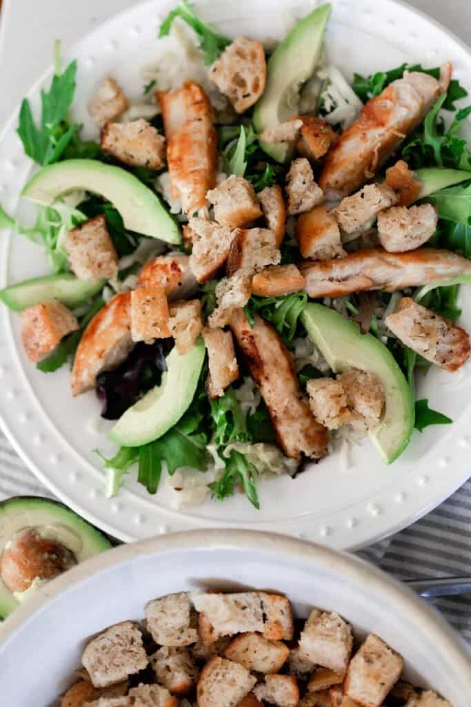 overhead photo of salad, avocado slices, and homemade croutons on a white plate with a bowl of croutons in the front