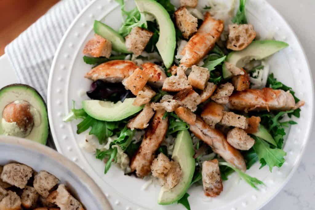 salad topped with croutons and avocado on a white plate on a stripped towel with a half of avocado and more croutons in a bowl on the side