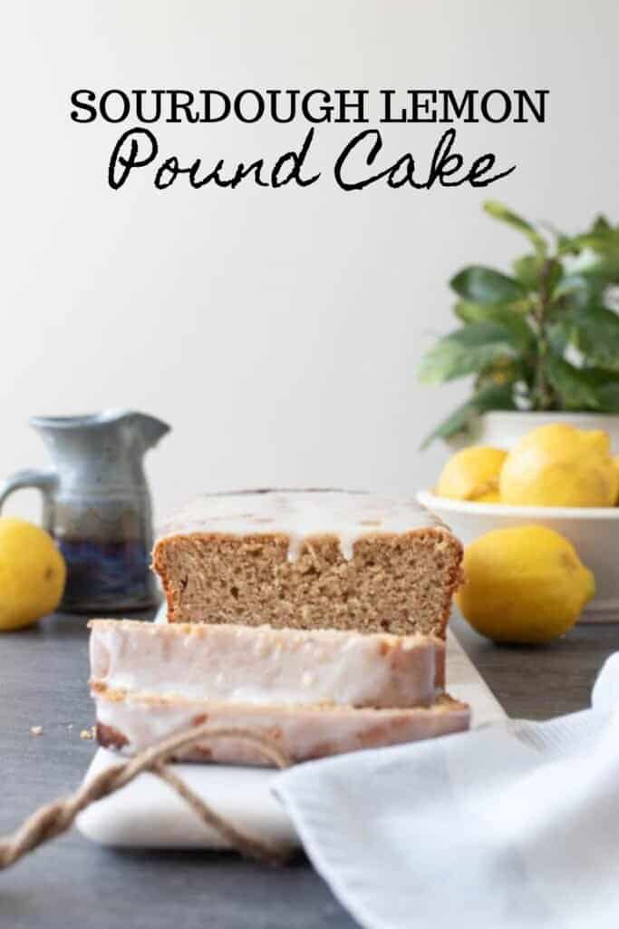 sourdough lemon pound cake with a lemon glaze sliced on a marble cutting board. A small gray pitcher, lemons, and a potted plant in the background