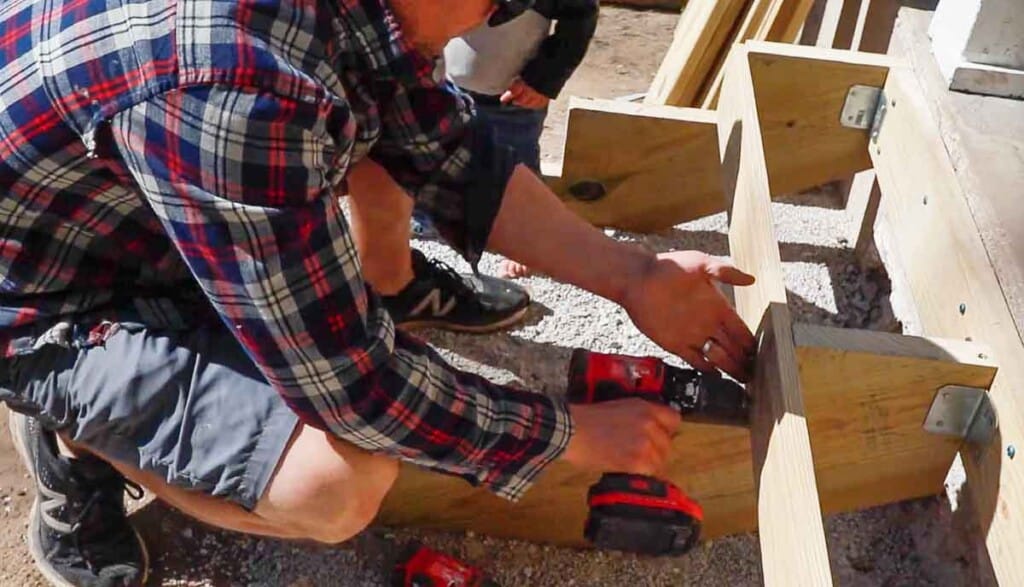 Man wearing a plaid shirt attaching risers to the front of stair stringers using a drill.