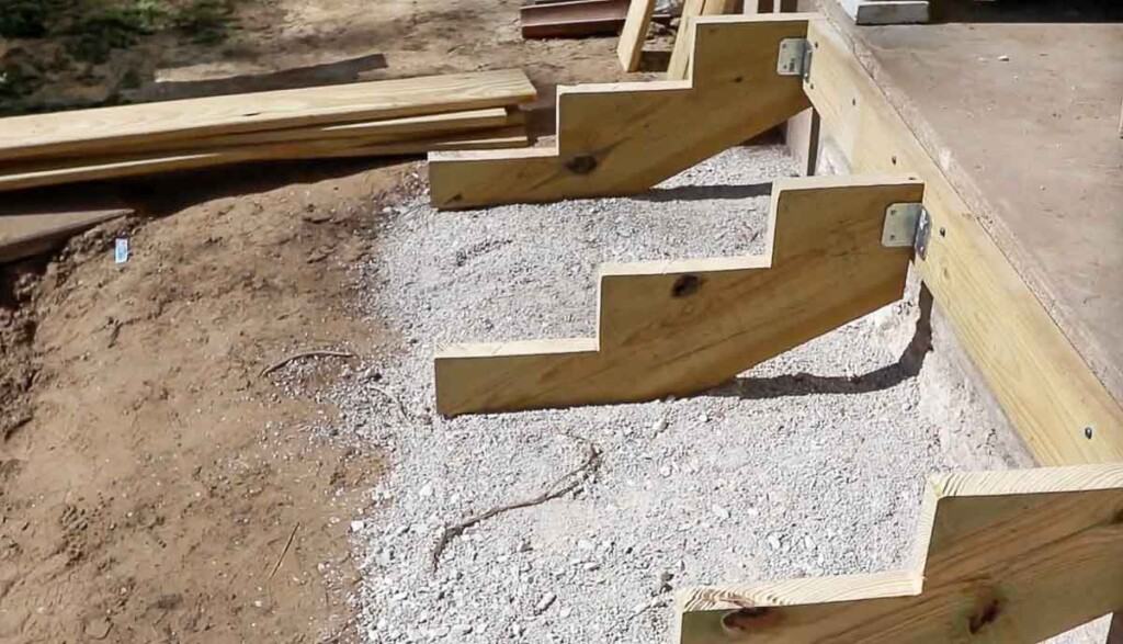 three stair stringers equally spaced attached to an anchor board with brackets. The stringers sit on a layer of gravel
