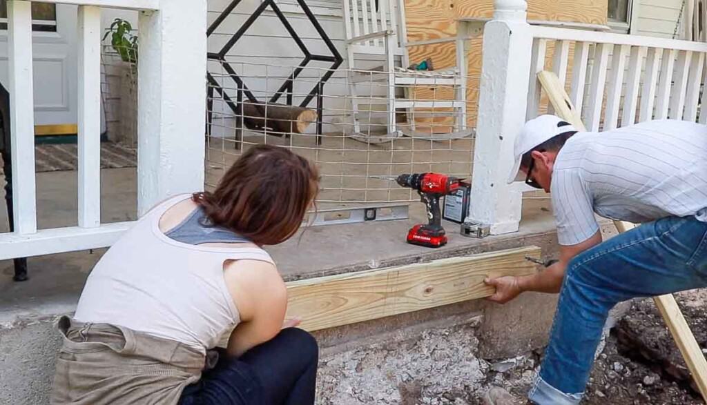 man and women drilling a wood board  to a concrete porch that will act as an an anchor for front steps.