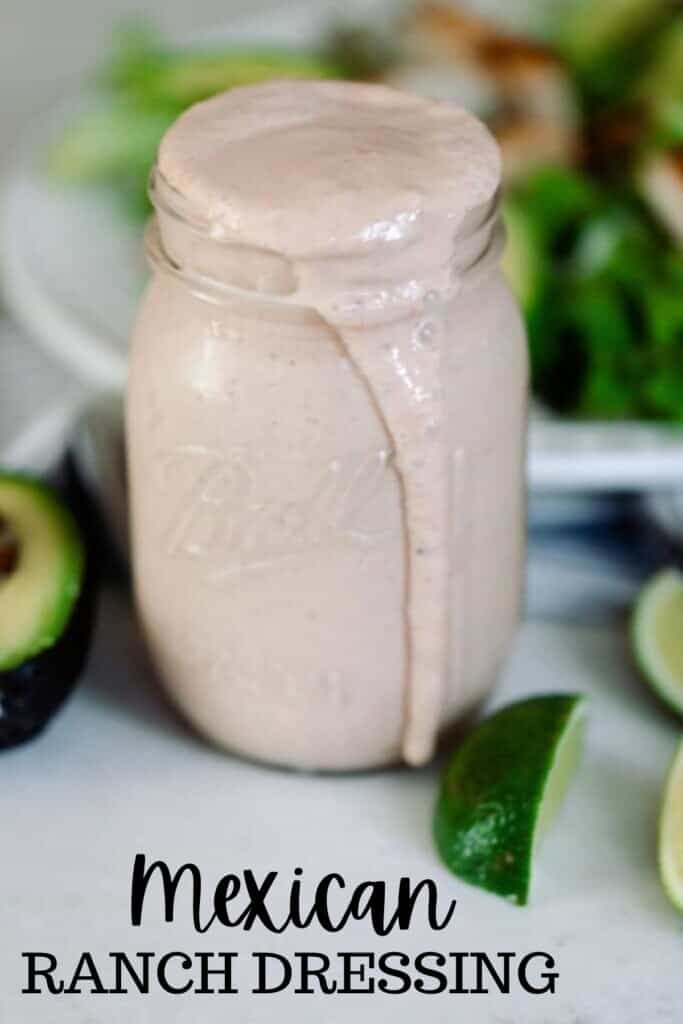 Mexican ranch dressing in a mason jar wit the dressing spilling over the top. Sliced limes and avocados lay on the counter around the jar of ranch