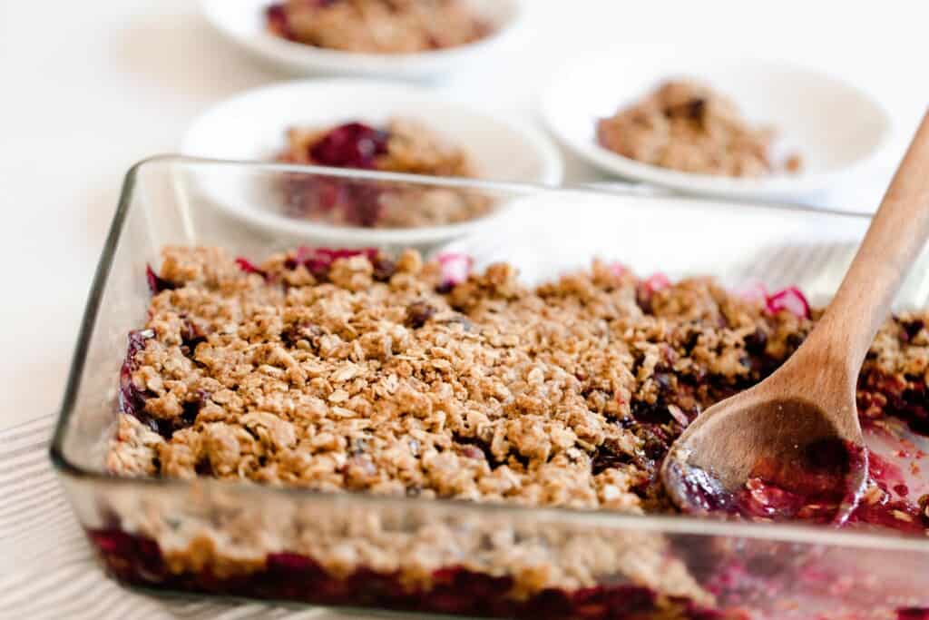 glass baking dish with einkorn berry crisp in it with a wooden spoon in the crisp. Plates of crisp are in the background