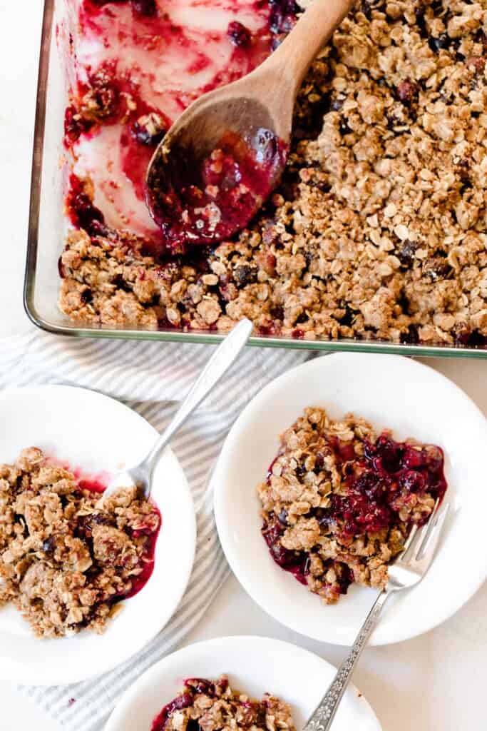 three white bowl of einkorn berry crisp with spoons with a glass baking dish of the remaining crisp in the background with a wooden spoon.