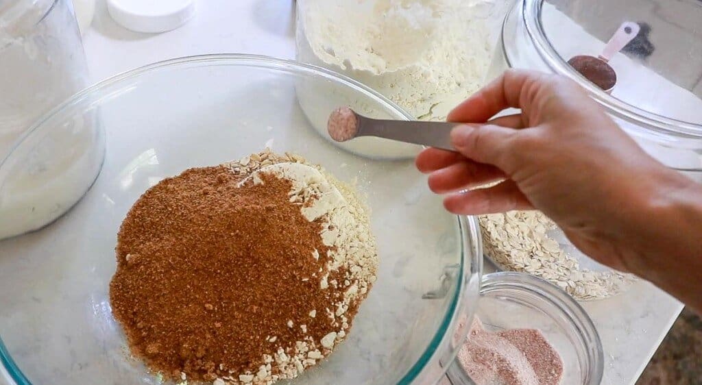 salt being added to a bowl of einkorn flour, coconut sugar, and oats in a glass bowl